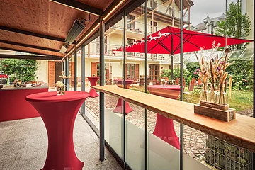 Überdachte Terrasse mit roten Stehtischen, Glaswand und Blick auf Garten mit rotem Sonnenschirm und Stühlen.