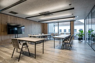 Moderner Besprechungsraum mit rechteckigen Tischen, Stühlen, großem Bildschirm und Fensterfront.