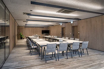 Konferenzraum mit U-förmigen Tischen, grauen Stühlen, Holzwänden und Deckenleuchten.
