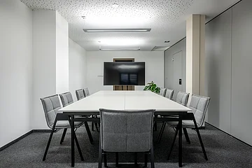 Konferenzraum mit weißem Tisch, zehn grauen Stühlen, Fernseher an der Wand und grauem Teppichboden.