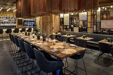 Moderner Gastraum mit Holztischen, schwarzen Stühlen, Kerzen und Pflanzen, Bar im Hintergrund.