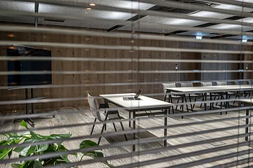 Blick durch Jalousien auf leeren Konferenzraum mit Tischen, Stühlen und offenem Laptop auf Tisch.