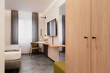 Hotelzimmer mit Bett, Schreibtisch, grünem Stuhl, Flachbildfernseher und Kleiderschrank aus Holz.