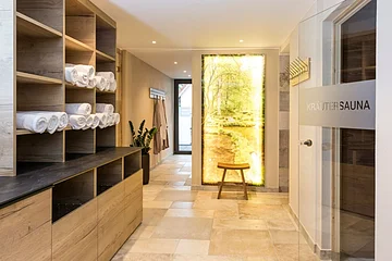 Flur mit Holzregalen und weißen Handtüchern, Tür mit Aufschrift „Kräutersauna“ und beleuchtetes Naturbild.