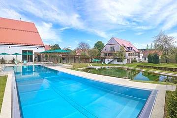 Außenbereich mit großem Schwimmbecken, Liegestühlen und Gebäuden mit roten Dächern bei blauem Himmel.