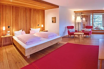 Schlafzimmer mit Holzverkleidung, Doppelbett, zwei roten Sesseln, Tisch, Stehlampe und großem Fenster mit Blick auf Terrasse.