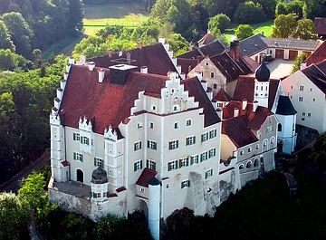 Luftaufnahme eines historischen Schlosses mit roten Dächern, weißen Mauern und umliegendem Wald.