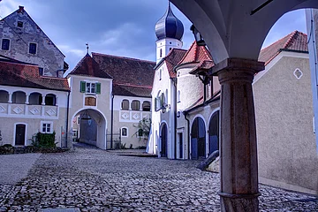 Innenhof eines historischen Gebäudekomplexes mit Kopfsteinpflaster und Torbogen unter bewölktem Himmel.