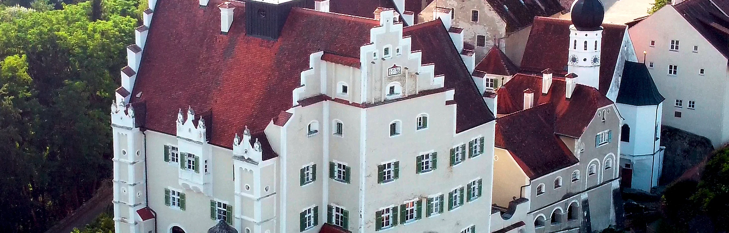 Luftaufnahme eines historischen Schlosses mit roten Dächern, weißen Mauern und umliegendem Wald.