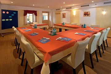 Konferenzraum mit U-förmiger Tischanordnung, orangen Tischdecken und weißen Stühlen, mit Flipchart und Fenstern.