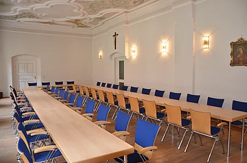 Konferenzraum mit langen Holztischen, blauen Stühlen, weißer Wand, Kreuz und Bild an der Wand, verziertem Deckenmuster