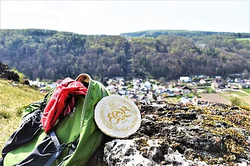 Grüner Rucksack mit rotem Tuch und rundem Bierdeckel auf Felsen, im Hintergrund Dorf und bewaldete Hügel.