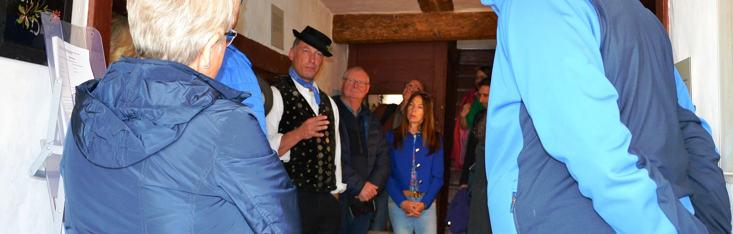 Gruppe von Menschen in einem engen Raum, einige hören einem Mann in traditioneller Kleidung zu.