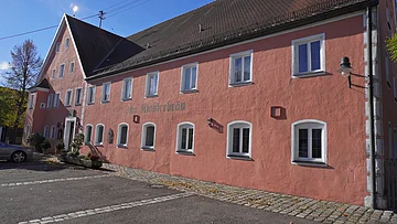 Rosa gestrichenes Gebäude mit weiß gerahmten Fenstern und Pflastersteinen davor bei Sonnenschein.