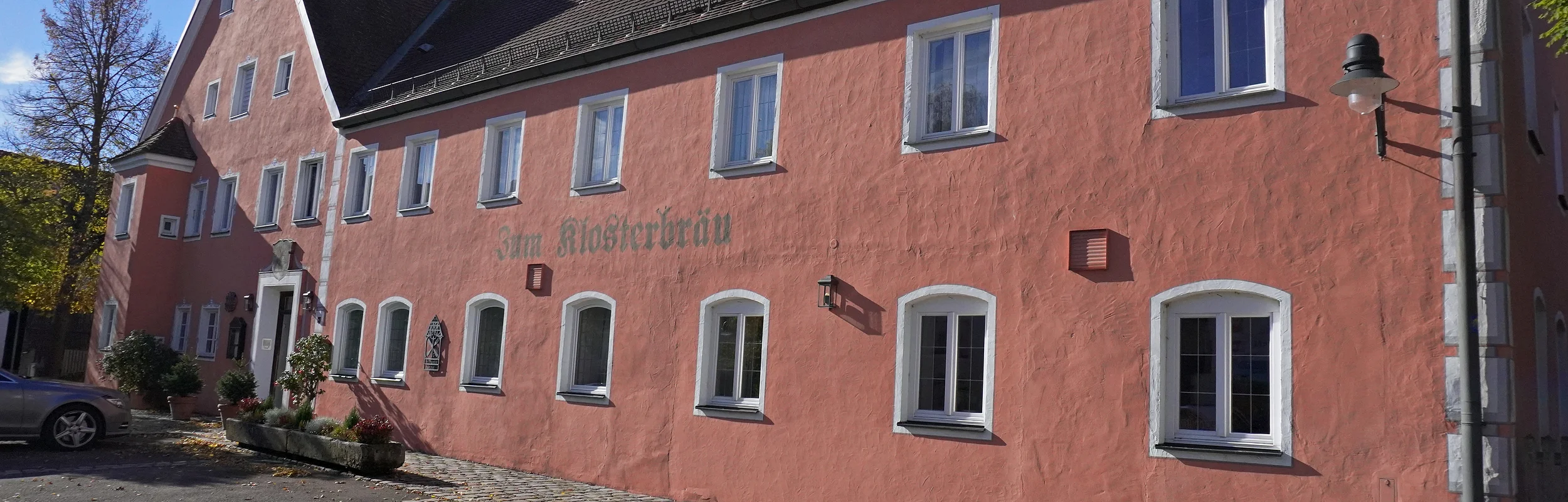 Rosa gestrichenes Gebäude mit weiß gerahmten Fenstern und Pflastersteinen davor bei Sonnenschein.