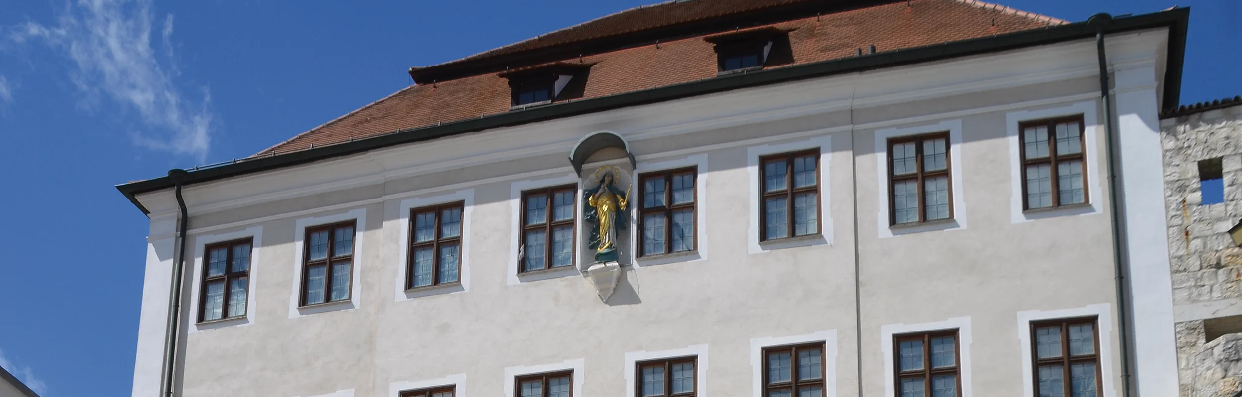 Wasserspiele vor einer historischen Fassade mit Statue und Text an einer Betonwand bei klarem Himmel.