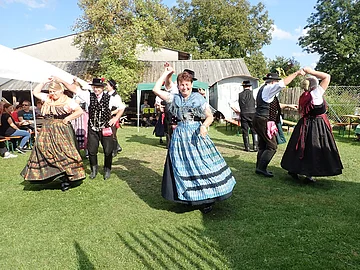 Drei Paare in Tracht tanzen im Garten des Jura-Bauernhof-Museum Hofstetten. Im Hintergrund ist ein Zelt und eine Scheune zu erahnen.