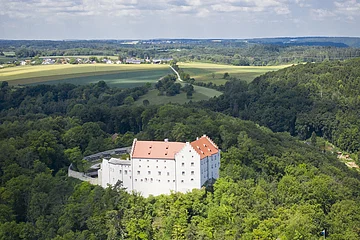Luftaufnahme eines weißen Schlosses mit rotem Dach, umgeben von Wald und Feldern unter blauem Himmel.
