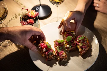 Zwei Hände nehmen kleine belegte Brotscheiben von einem weißen Teller auf einem Holztisch.