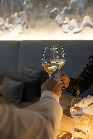 Zwei Personen stoßen mit Weißweingläsern an einem Holztisch in einem Restaurant an.
