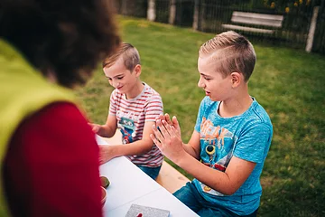 Zwei Jungen sitzen an einem Tisch im Freien, ein Erwachsener ist unscharf im Vordergrund zu sehen.
