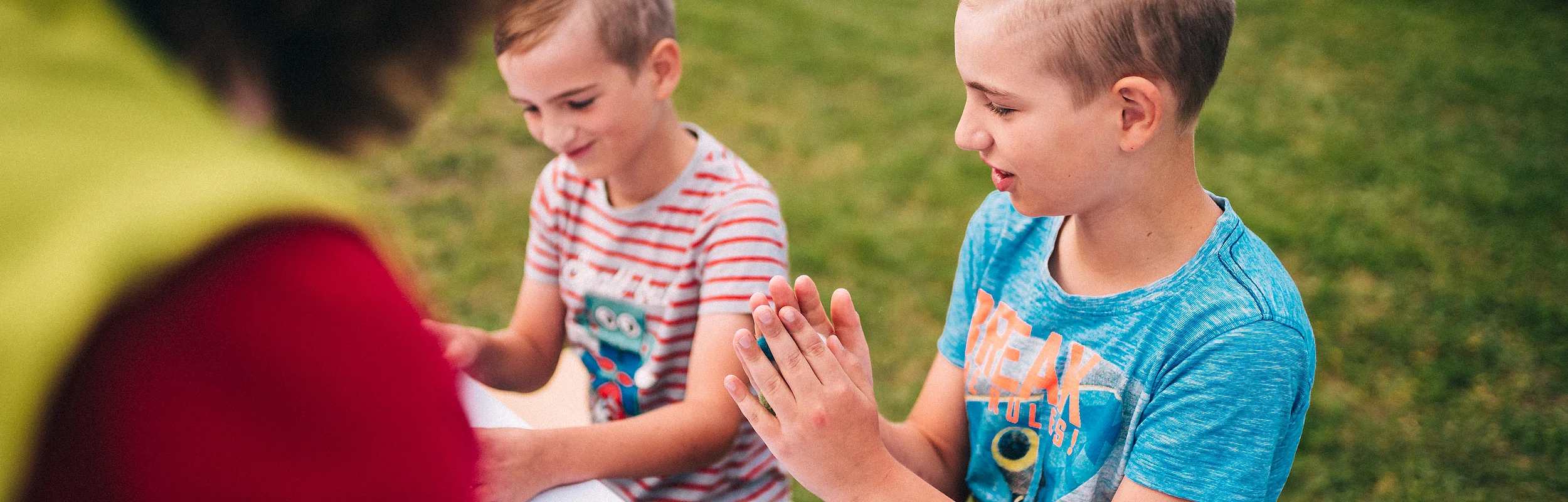 Zwei Jungen sitzen an einem Tisch im Freien, ein Erwachsener ist unscharf im Vordergrund zu sehen.