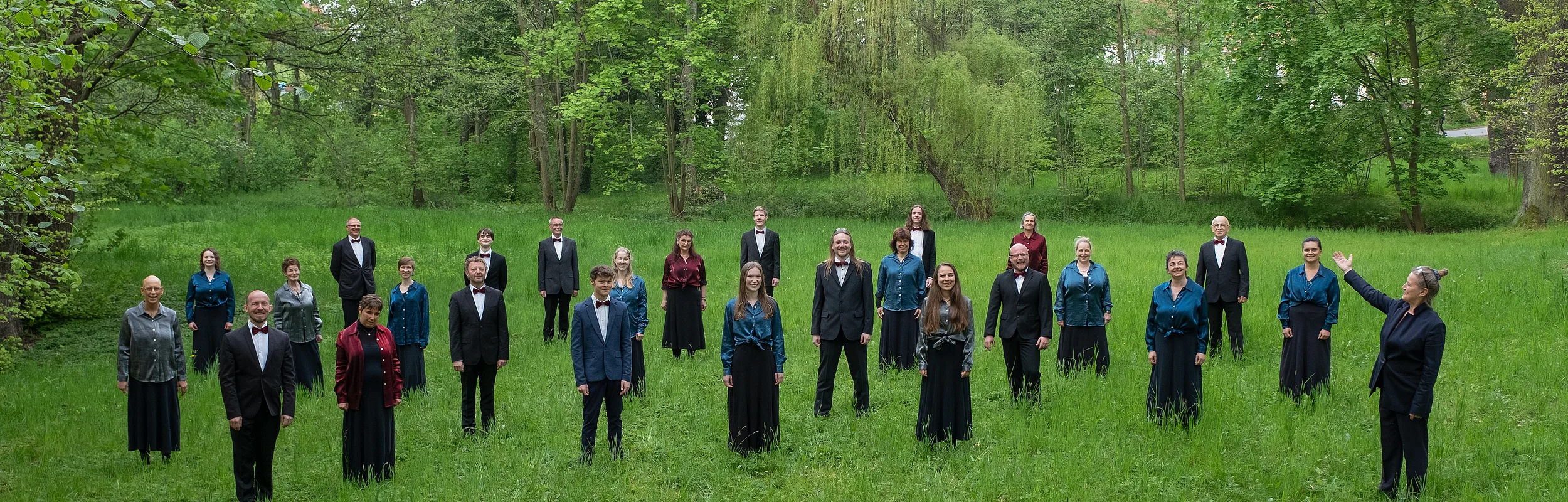 Gruppenfoto von 24 Personen in formeller Kleidung auf einer Wiese vor Bäumen, rechts vorne eine Person winkt.