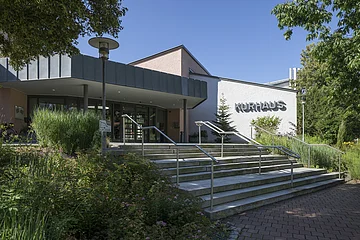 Eingang eines Gebäudes mit Treppen, Handläufen und dem Schriftzug „Kurhaus“ an der weißen Fassade.