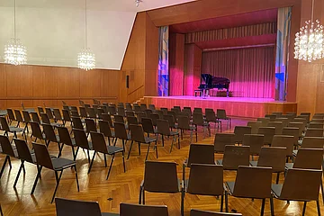 Leerer Konzertsaal mit Reihen von Stühlen und einem Flügel auf der Bühne mit roter Beleuchtung.