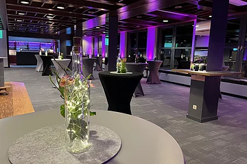 Leerer Veranstaltungsraum mit Stehtischen, Flaschen mit Lichterketten und violetter Wandbeleuchtung.