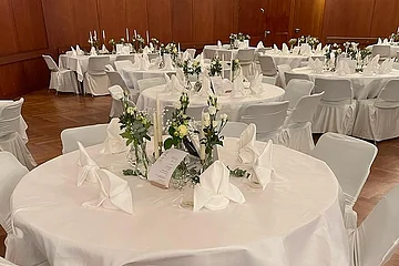 Rund gedeckter Tisch mit weißen Hussen, Servietten, Kerzen und Blumen in einem Saal mit Holzwänden.