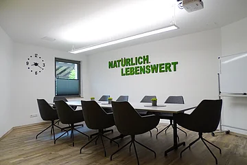Konferenzraum mit Tisch, acht Stühlen, Uhr, Fenster, Whiteboard und Schriftzug „Natürlich. Lebenswert“ an der Wand.