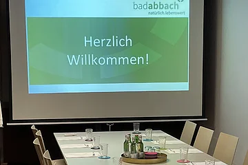 Konferenzraum mit langem Tisch, Stühlen, Gläsern, Flaschen und Projektionsbild mit Text „Herzlich Willkommen!“