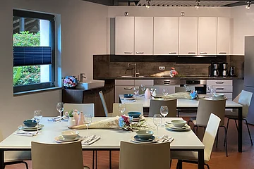 Küche mit zwei gedeckten Tischen, weißen Schränken, Spüle, Kaffeemaschinen und Fenster mit Blick ins Grüne.