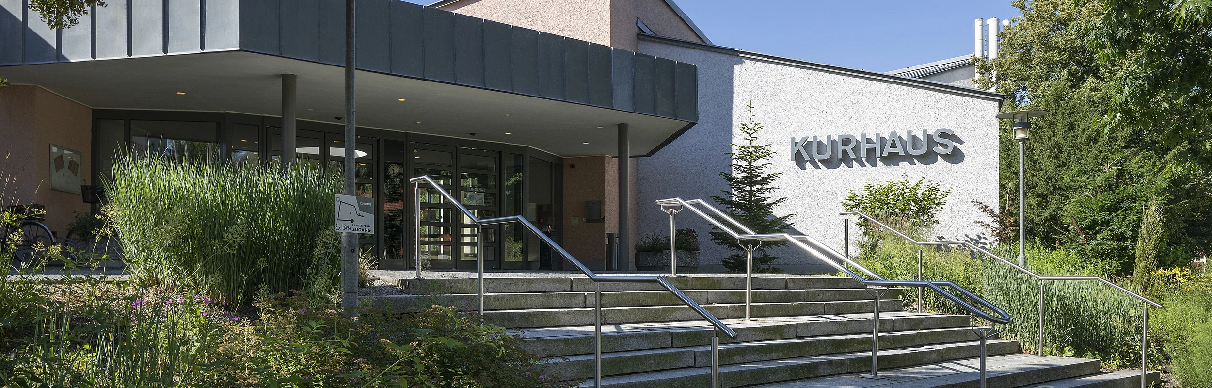 Eingang eines Gebäudes mit Treppen, Handläufen und dem Schriftzug „Kurhaus“ an der weißen Fassade.