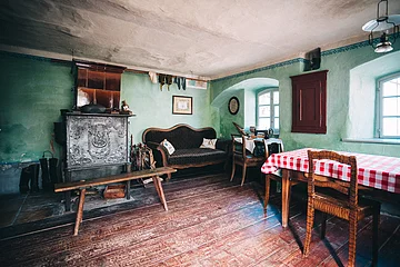 Rustikaler Raum mit grün gestrichenen Wänden, einem Ofen, einer Couch, einem Holztisch mit rot-weiß kariertem Tuch und Stühlen.
