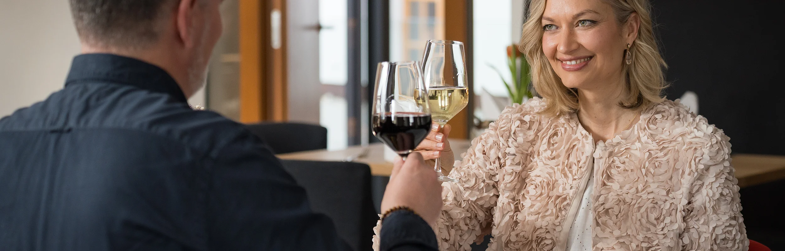 Paar sitzt in Restaurant und stößt mit Rot- und Weißweinglas an, Frau lächelt.