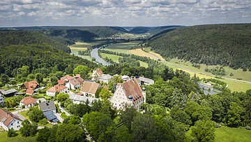 Luftaufnahme eines Dorfes mit Fluss, umgeben von bewaldeten Hügeln und Feldern unter bewölktem Himmel.