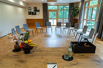 Seminarraum mit Stühlen im Halbkreis, großem Fenster, Holzboden, Tisch mit Obst, Wasserflasche und Notizblock.