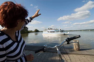 Eine Fahrradfahrerin winkt vom Ufer aus der MS Altmühlsee zu.